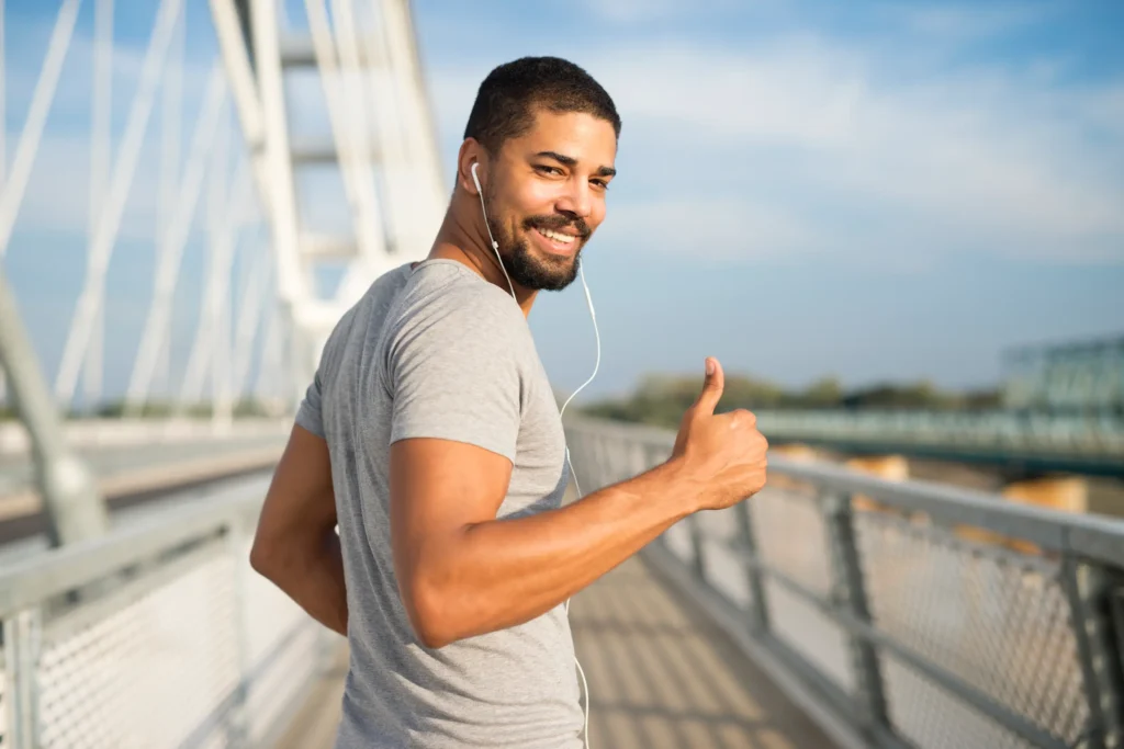Homem sorrindo enquanto caminha ao ar livre, com fones de ouvido, em uma ponte.