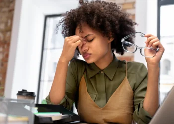 Mulher com expressão de cansaço e dor, segurando os óculos enquanto esfrega a testa, refletindo sobre os sinais de estresse mental.