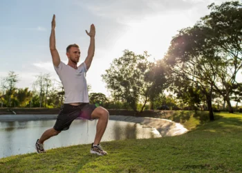 Homem praticando exercício de alongamento ao ar livre, com postura de lunge e braços elevados, em um ambiente natural.