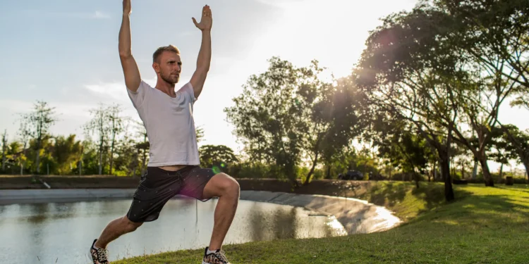 Homem praticando exercício de alongamento ao ar livre, com postura de lunge e braços elevados, em um ambiente natural.