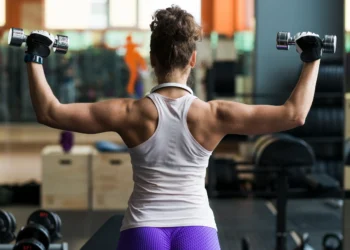 Mulher treinando ombro com halteres em academia para aumentar força e definição muscular.