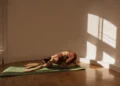 Praticante de yoga em casa realizando a postura da Criança (Balasana), com braços estendidos no tapete e iluminada suavemente pela luz natural.